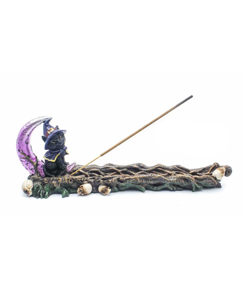 A gothic-themed incense holder featuring a small black cat in a pointed hat sitting on a purple crescent moon, with a long brown incense stick resting diagonally across it, all set on a gnarled tree branch base with small skulls.